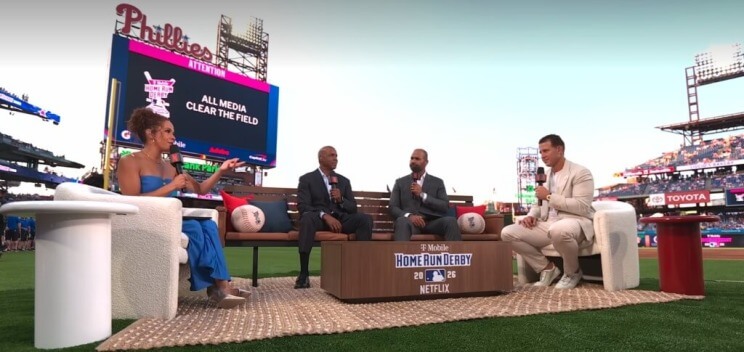 Elle Duncan, Barry Bonds, Albert Pujols, and Anthony Rizzo sit on couches set up on a baseball field. Behind them, a scoreboard message reads "ATTENTION: ALL MEDIA CLEAR THE FIELD"