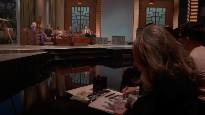 A courtroom sketch artist in the foreground drawing the people on John Mulaney's show seated on the couch in the background